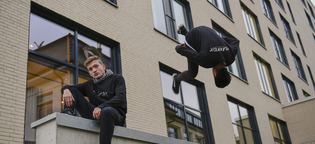 One Man sitting in front of a building, an other doing a Backflip down from a roof